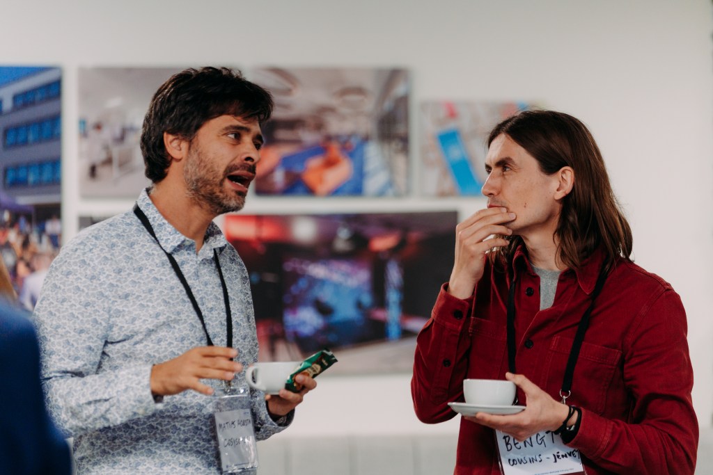 Two men having a conversation at Scale Space with coffee cups in their hand with a backdrop of art. Matias Acosta, CEO, Cosysense at the Better Futures Retrofit Accelerator's Challenge Identification Workshop. Photo by Velvet Green Media.